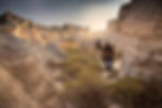 Two hikers walk through a rocky canyon carrying equipment. The sun sets behind them, illuminating the dramatic cliffs and sparse vegetation.