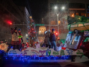 Parade float with people waving, colorful lights, and signs. Snowfall creates a festive atmosphere. Kansas Ave sign in background.