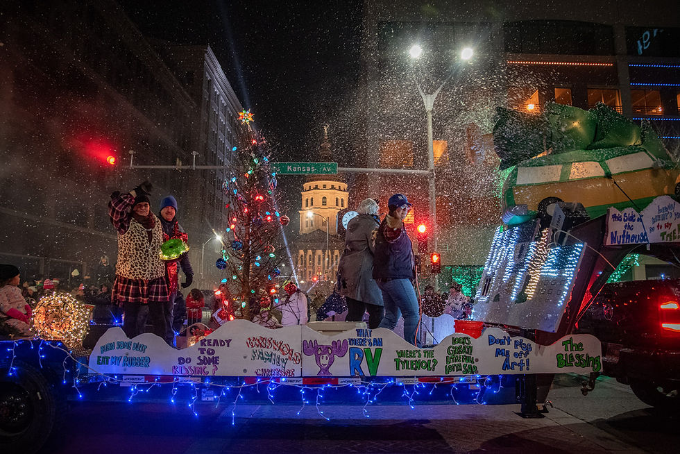 Parade float with people waving, colorful lights, and signs. Snowfall creates a festive atmosphere. Kansas Ave sign in background.