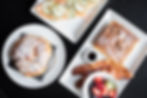 Pastry with powdered sugar, smoked salmon toast, and French toast with bacon and syrup on white plates, set on a dark table.