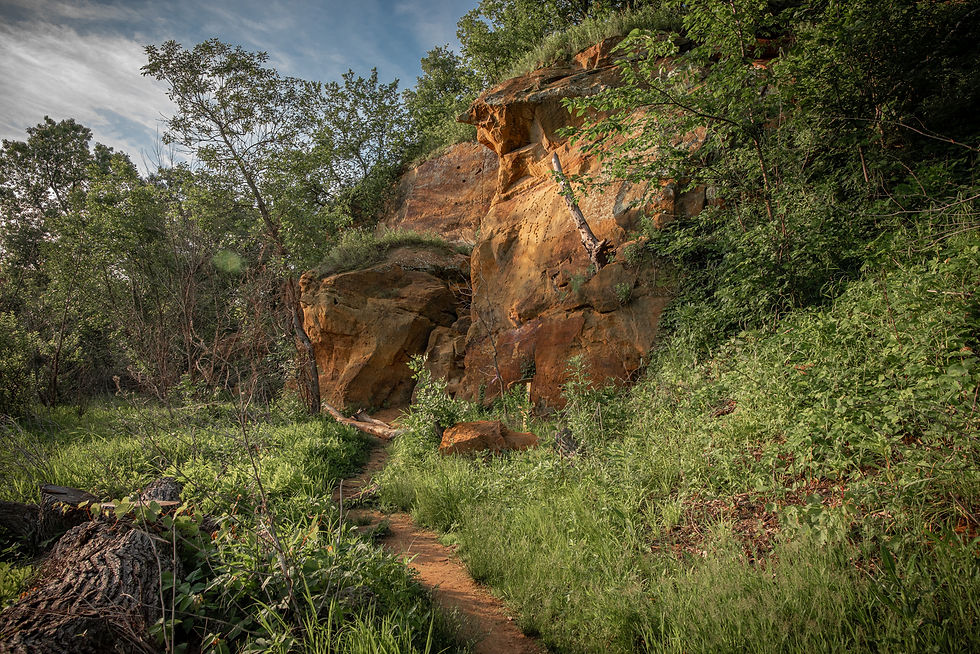 Dirt trail winding through lush green woods beside an orange sandstone cliff under a partly cloudy sky.