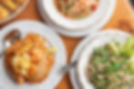 Plates of Thai food on a wooden table include Pad Thai with shrimp, a spicy salad, and dumplings. Natural light highlights the vibrant colors.