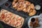 Pizza with chicken and tomatoes, waffle fries with sauce, and fried balls with dip on black trays; appetizing and colorful setting.