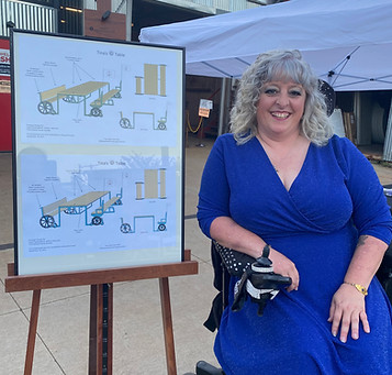 A smiling woman in a blue dress sits in her wheelchair next to an easel with a sketch of the new Tina’s Table adaptive seating