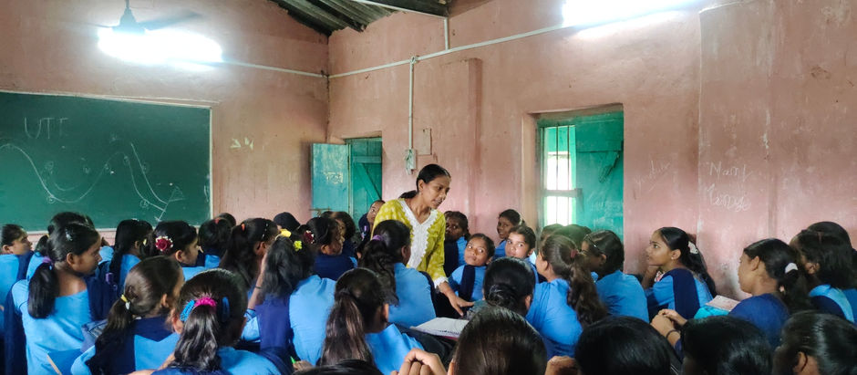 ZealGrit's facilitator Sandhya in yellow engages with students in blue uniforms in a classroom. Chalkboard with diagrams in background. Bright ambiance.