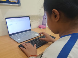 Suhani in white and blue shirt types on a laptop at a wooden desk. Posters are on the wall, and a purple bottle sits nearby.