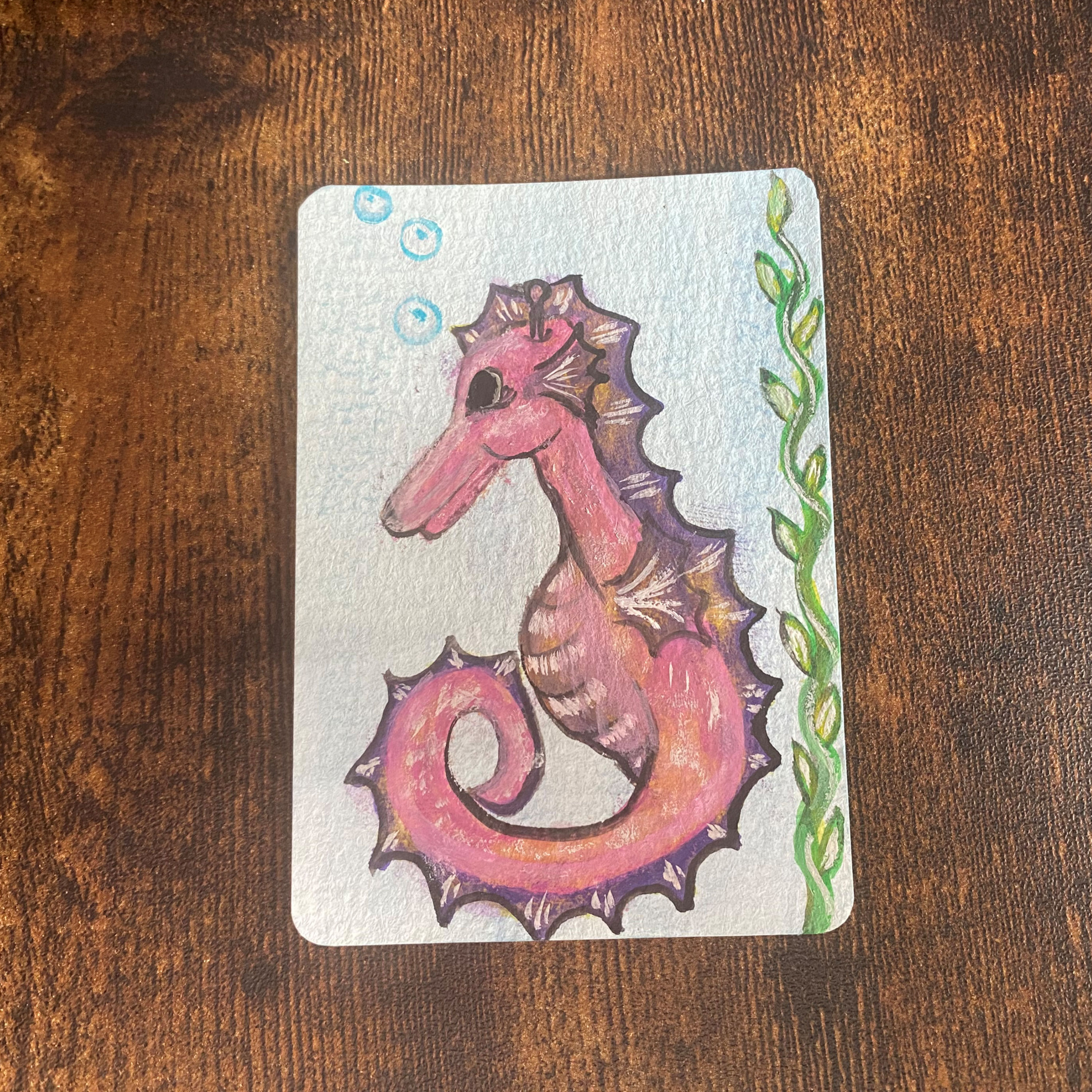 hand drawn watercolour painting of a pink and purple seahorse with green seaweed top to bottom right hand side