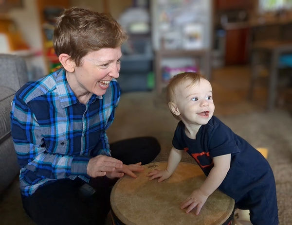 An adult and a baby are sitting on the floor, playing a large gathering drum with their hands. The adult has short, light brown hair and is wearing a blue flannel shirt. They are looking at the baby and smiling. The baby has a small amount of blonde hair and is looking away, smiling.