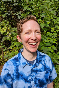 A light skinned adult with short, wavy, dark blond hair is smiling at the camera outside in front of a green bush. They are wearing a blue tie dye shirt.