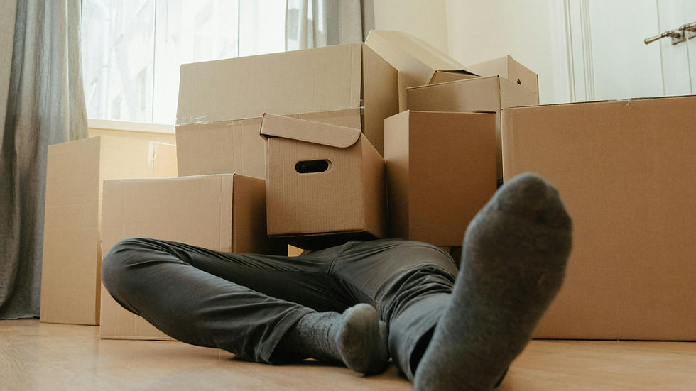 Person lying under stacked cardboard boxes in a bright room with wooden floor, wearing dark pants and socks. A sense of exhaustion is conveyed.