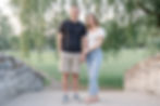 An engaged couple holds hands on a stone bridge in Upper Onondaga park in Syracuse, NY. The man wears a black shirt and khaki shorts, the woman in a white top and jeans. Greenery surrounds them.
