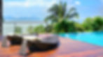Two wicker loungers with cushions on a wooden deck beside an infinity pool, overlooking a tropical landscape with palm trees and distant mountains.