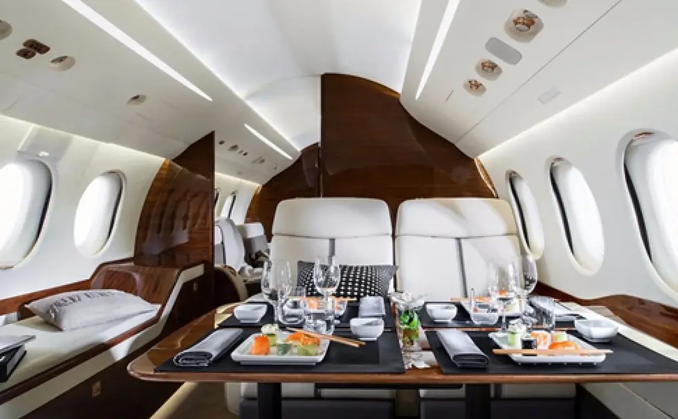 Luxurious private jet interior with white leather seats, wood accents, and a dining table set elegantly with sushi and wine glasses.