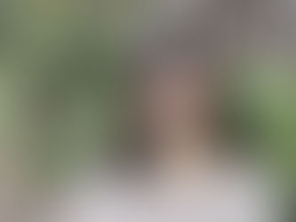 A woman with dark hair smiles broadly in an outdoor setting with greenery. She wears a light sweater, creating a cheerful mood.