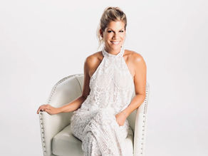 Woman in a white lace dress seated on a white studded chair, smiling against a plain white background.