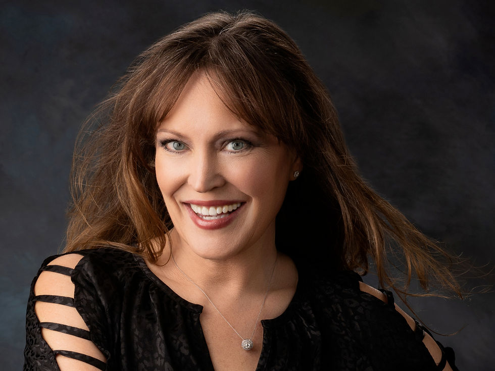 Smiling woman with brown hair wearing a black patterned top and necklace. Dark, blurred background; relaxed and cheerful mood.