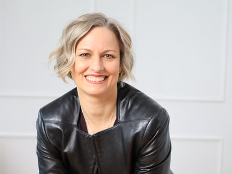 Smiling blonde woman in a black leather jacket poses against a white wall, with a bright, friendly expression.