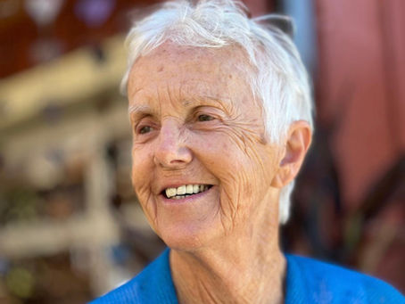 Lori Pappas with short white hair smiling warmly. Wearing a blue garment with a blurred outdoor background and reddish hues.