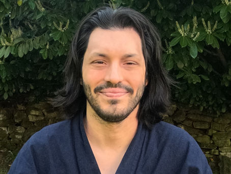 Man with long hair and a navy shirt smiles softly outdoors, set against lush green foliage and stone wall background. Calm, serene mood.