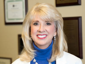 Diana Driscoll with blonde hair, wearing a blue turtleneck and white coat, in an office with framed certificates on the wall.