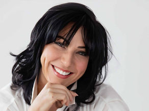Smiling dark-haired woman in a white shirt, resting her chin on her hand against a plain white background.