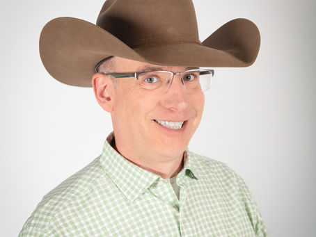 Man wearing glasses and a brown cowboy hat smiles against a plain white background. He has a green checkered shirt, creating a joyful mood.