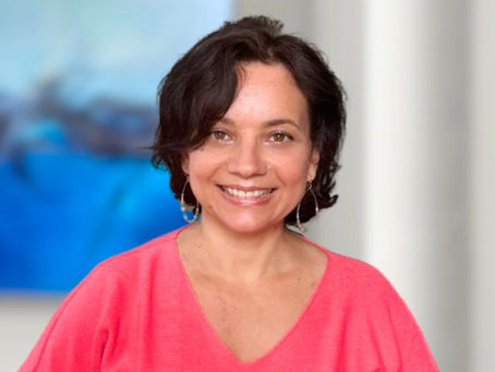 Smiling woman in a pink top stands indoors, with a blurred blue abstract painting in the background. Bright and cheerful mood.