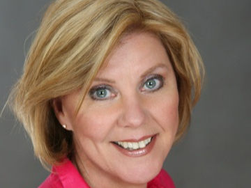 Smiling woman with blonde hair in a pink shirt against a gray background, conveying a cheerful mood.