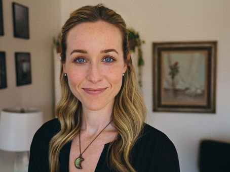 Smiling woman with long blonde hair in a home setting. She's wearing a necklace, with framed art and plants in the background. Calm mood.