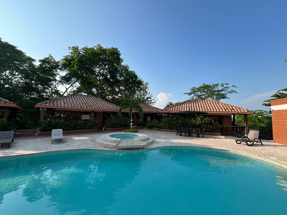 This image shows a serene poolside area at what appears to be a resort or vacation property. The crystal-clear pool is surrounded by stone-tiled decking, lounge chairs, and a circular jacuzzi at the center. In the background, several cozy cabanas or bungalows with terracotta-tiled roofs are nestled among lush tropical greenery, creating a relaxing and inviting environment. The open, bright sky and peaceful ambiance make this setting ideal for relaxation and leisure under the warm sun
