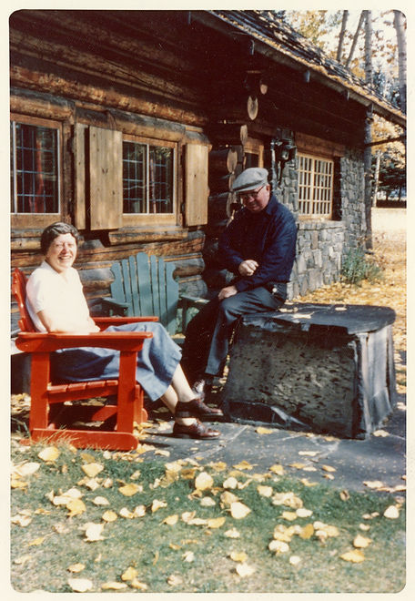 [Catharine and Peter Whyte seated outside their home in Banff]. [ca.1963]. Peter and Catharine Whyte fonds. V683 / III / A / 1 / PA - 42. An older, smiling Catharine and Peter Whyte sitting outside the front of their home, Catharine in a red wooden chair and Peter on the stone slab, with yellow leaves on the ground in colour.  Inscription on the back: “Katherine + Peter Whyte at their home in Banff Alberta, Canada 1960 Photo by: Miss Christine L. Reid 86 Buckinham Street, #24 Cambridge Mass. 02138 - U.S.A.”