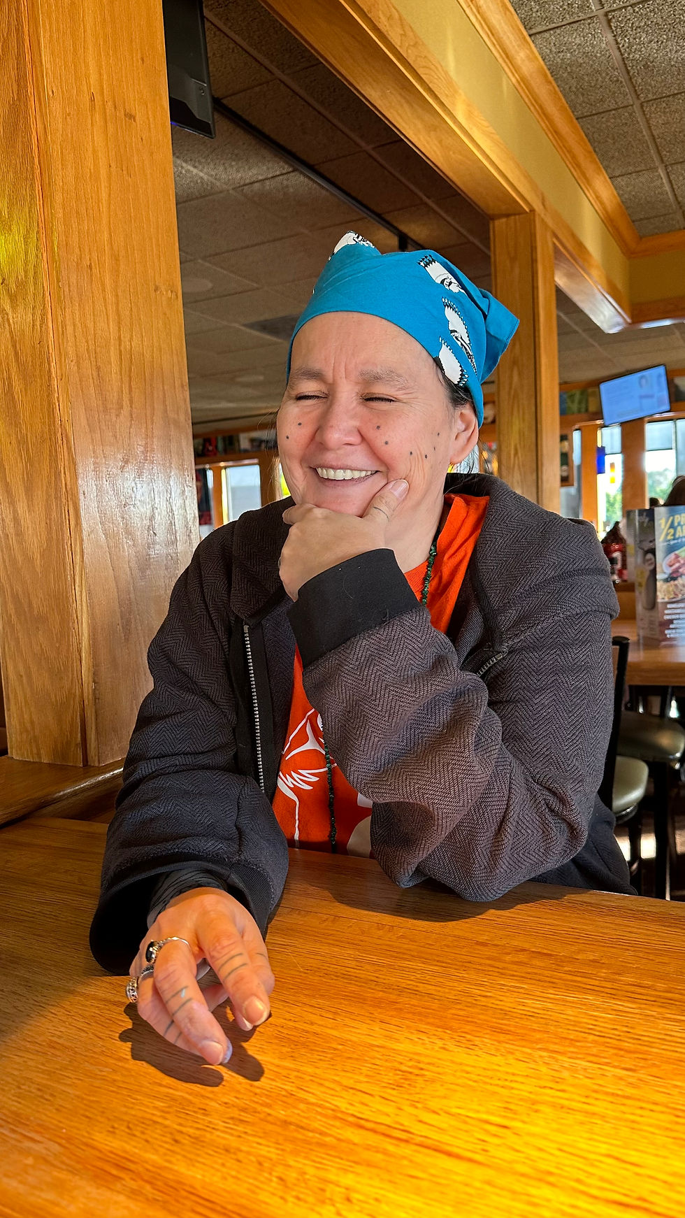 The artist Ella Nathanael Alkiewicz sits at table with smile and eyes closed. Elbows on table and she wears a gray sweatshirt, orange tshirt and blue bandana on her head. 