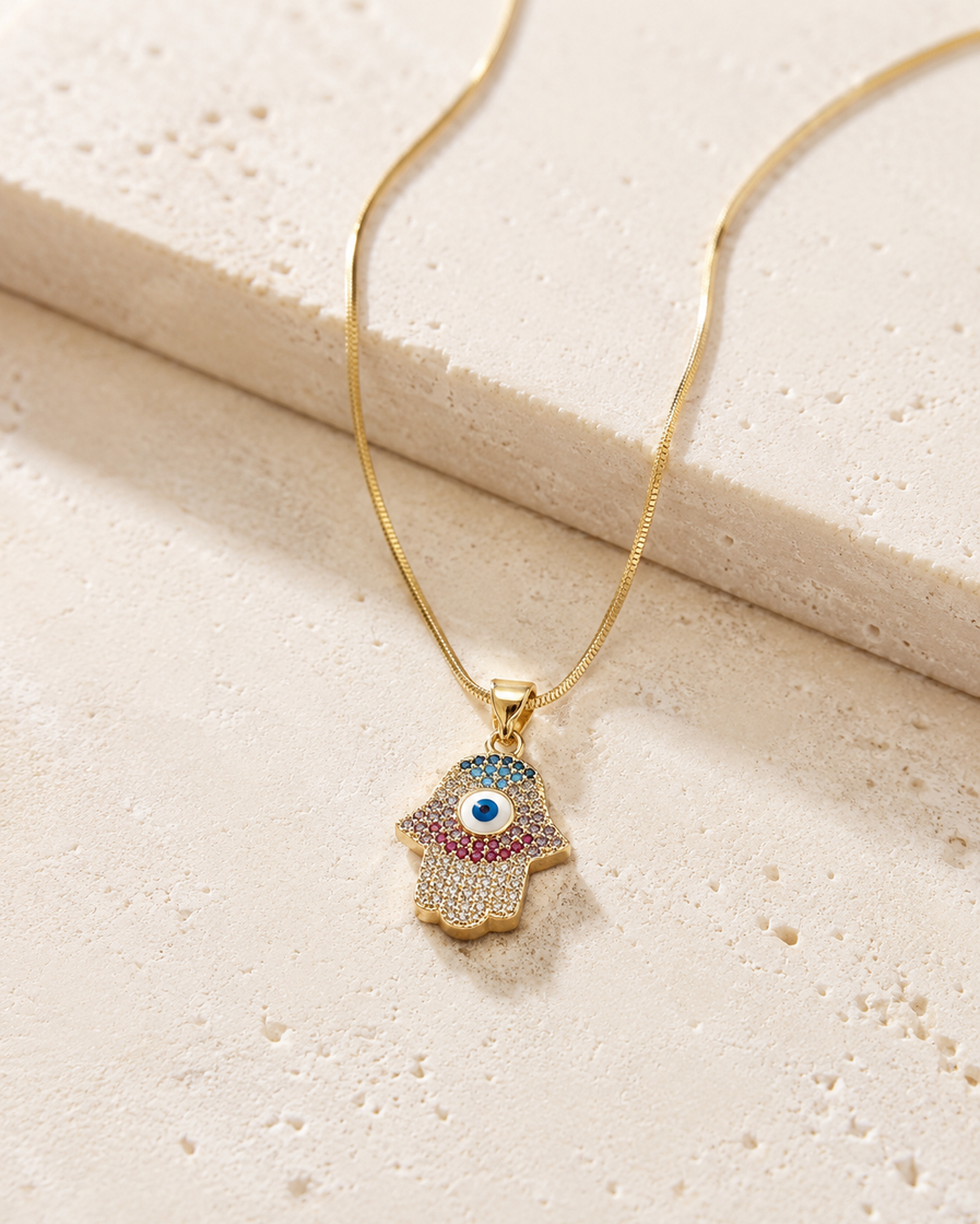 Gold-tone Hamsa evil eye pendant necklace with multicolour crystals on a cream stone surface