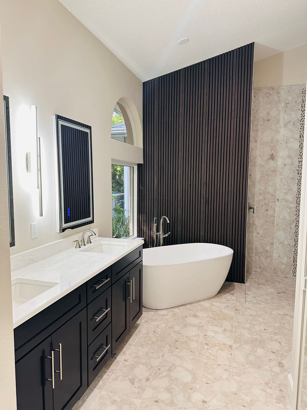 Modern bathroom with a freestanding tub, dark wooden accent wall, double sink vanity with black cabinets, marble floor, and arched window.