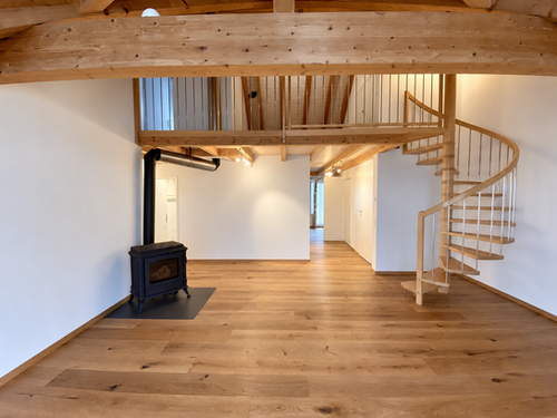 Wohnraum mit Holzparkett, sichtbaren Deckenbalken, Galerie, Wendeltreppe und schwarzem Schwedenofen.