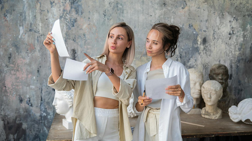 Two women in an art studio discussing and analyzing sculptures, with papers in hand.