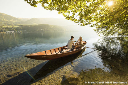 Paar im Ruderboot auf hellem See, Berge, Sonne, Äste im Vordergrund.