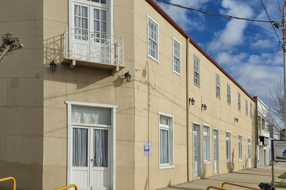 Fotografía del edificio del ex Hotel Colón en Puerto San Julián, actualmente Residencia Estudiantil. La fachada de estilo magallánico, en tonos ocres, presenta una esquina destacada con un balcón en el primer piso y grandes ventanales con marcos blancos. Se observan detalles como faroles antiguos y barandas amarillas que delimitan la acera