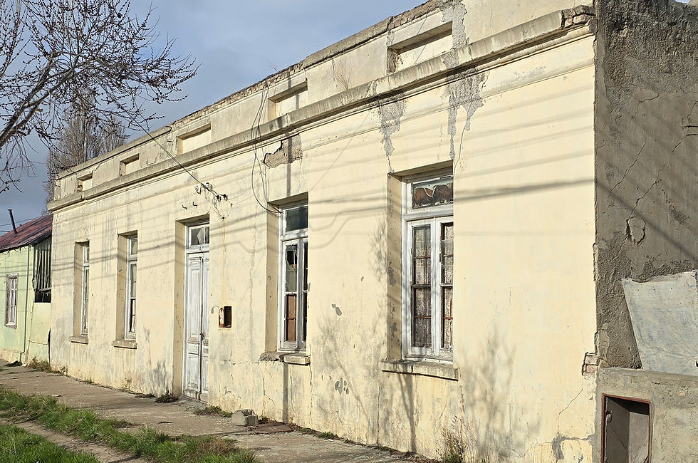 Fotografía de la fachada en ruinas de la ex comisaría en Puerto San Julián, declarada Monumento Histórico Nacional. El edificio de una planta, de color amarillo pálido desgastado, muestra signos de deterioro con grietas y pintura desgastada. Se aprecian ventanas con marcos de madera y vidrios rotos, así como una puerta principal vieja de madera. Un árbol sin hojas se encuentra a la izquierda, y la vereda frente al edificio está descuidada.