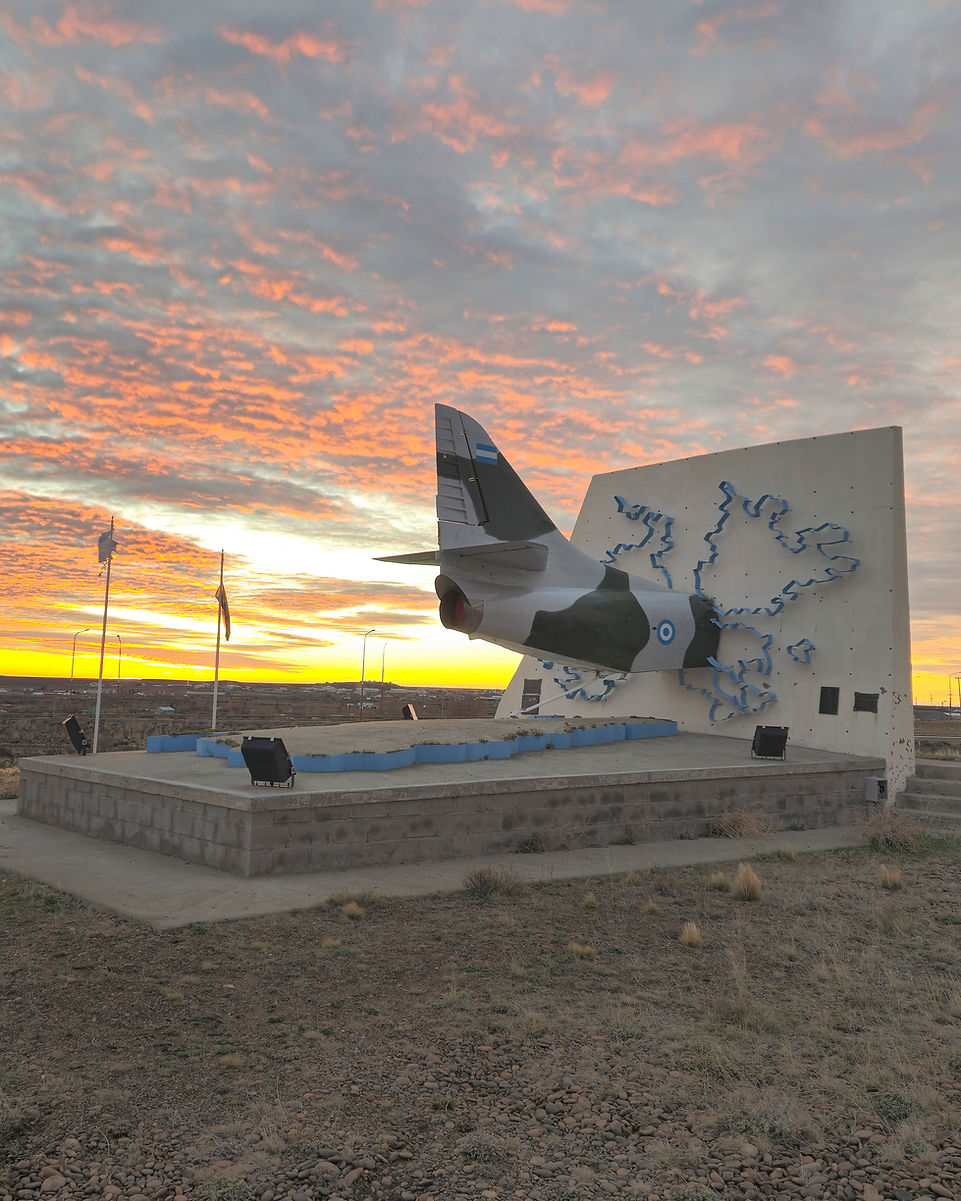 Fotografía de un monumento en las afueras de Puerto San Julián. La escultura principal muestra la cola de un avión de combate incrustada en un muro donde está un mapa en relieve de las Islas Malvinas. El monumento se alza contra un cielo anaranjado al amanecer.