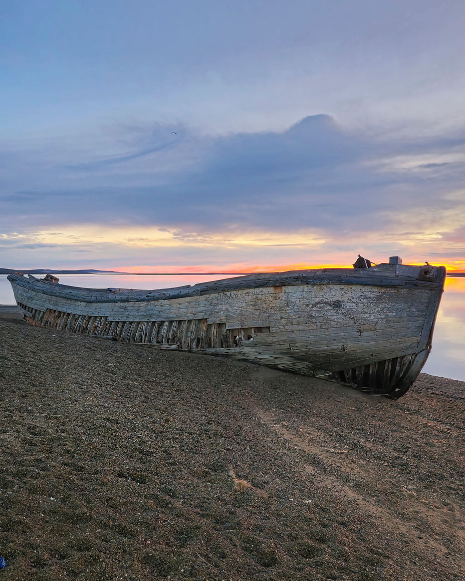 Una vieja barcaza de madera encallada en la orilla la bahía al amanecer. La embarcación está inclinada hacia un lado, con las costillas expuestas y la madera desgastada por el tiempo. El cielo está pintado con colores vibrantes del atardecer, reflejándose en el agua tranquila.