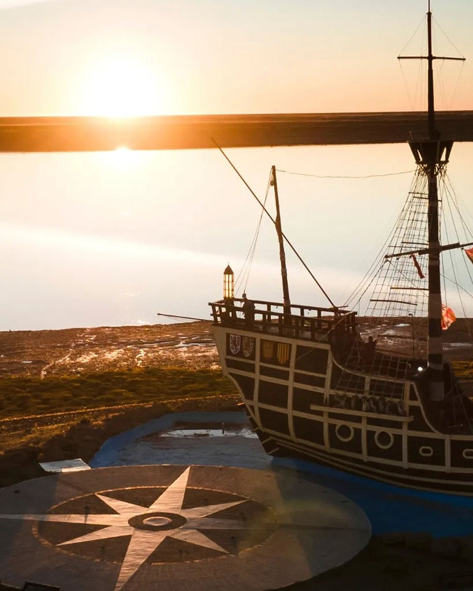 Una réplica de la Nao Victoria, el primer barco en circunnavegar el mundo, se alza majestuosa en primer plano contra un impresionante atardecer. El sol poniente proyecta un resplandor dorado sobre el barco y el paisaje circundante. En el suelo, justo debajo de la proa, se encuentra el Hito Punto Cero, una rosa de los vientos que marca el inicio la patagonia.