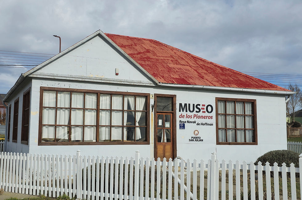 Fotografía de una antigua casa de una sola planta con techo rojo brillante y varias ventanas, detrás de una cerca blanca de madera. Un cartel sobre la entrada dice "Museo de los Pioneros Rosa Novak de Hoffman"