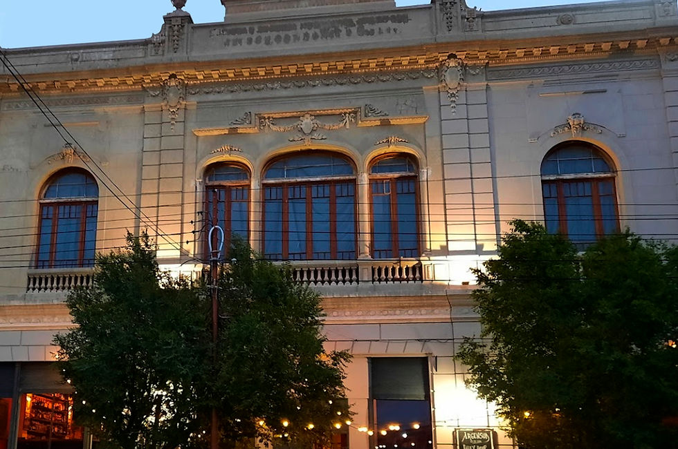 Imponente fachada del edificio Argensud al atardecer. La estructura histórica exhibe una arquitectura elegante con detalles ornamentales en su parte superior y grandes ventanales arqueados en el primer piso. Luces cálidas iluminan la fachada y los árboles a ambos lados. Un automóvil compacto y una camioneta estacionados en la calle frente al edificio añaden un toque de vida cotidiana a la escena.