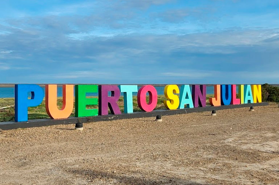 Grandes letras coloridas escritas "PUERTO SAN JULIAN" aparecem sobre uma paisagem costeira. O céu é azul brilhante com algumas nuvens, e você pode ver a água da baía ao longe.