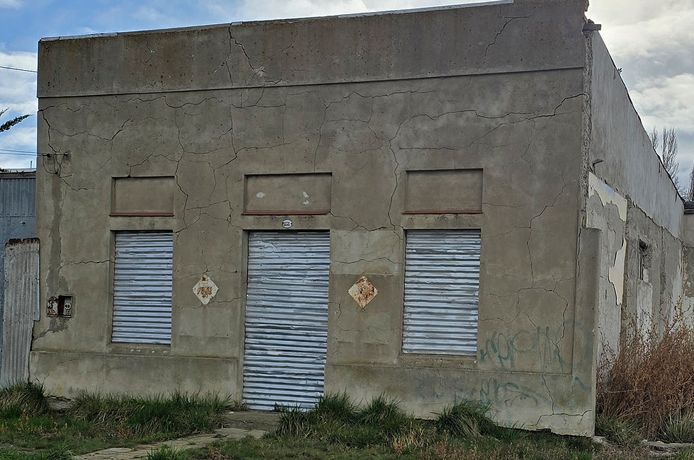 Fotografía de la fachada deteriorada del ex prostíbulo 'La Catalana' en Puerto San Julián. El edificio de una planta, con paredes de cemento agrietadas y descoloridas, muestra signos evidentes de abandono. Tres grandes persianas metálicas cerradas ocupan la mayor parte de la fachada. Se ve maleza creciendo frente a la fachada.