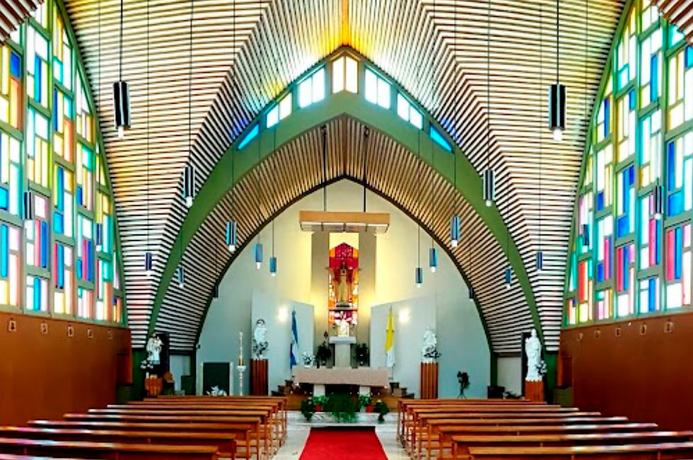 fotografia interior de la Parroquia del Sagrado Corazón de Jesús en Puerto San Julián. El techo abovedado de madera con vigas a la vista crea un efecto visual llamativo. A ambos lados, vitrales coloridos con diseños geométricos iluminan el espacio. En el fondo, el altar destaca con una alfombra roja que lo conduce, y una imagen religiosa en el centro. Filas de bancos de madera se extienden a lo largo de la nave central, invitando a la oración y la reflexión.