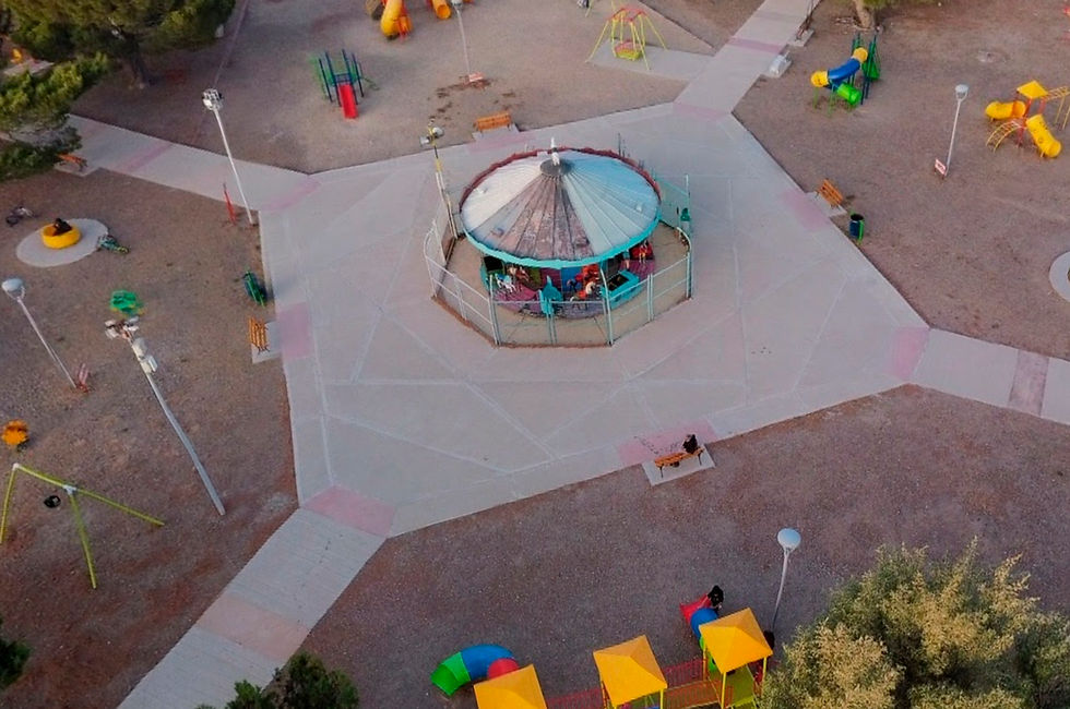 Vista aérea de una plaza en Puerto San Julián. Un carrusel colorido ocupa el centro, rodeado de diversas áreas de juego con toboganes y estructuras para trepar. Senderos de concreto cruzan la plaza, bordeada de árboles y arbustos verdes. Algunas personas disfrutan del espacio al aire libre en este día soleado.