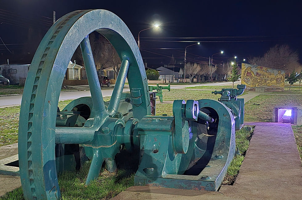 Fotografia de una gran pieza de maquinaria de color verde oscuro, probablemente una rueda dentada y parte de un motor, se alza en una plazoleta temática. Iluminada por la luz nocturna, esta reliquia industrial contrasta con las farolas y la tranquila calle de Puerto San Julián. Es un recordatorio del pasado del frigorífico Swift en la región