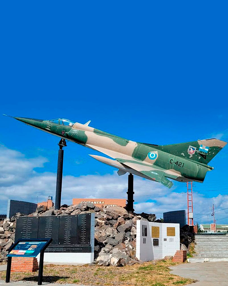 Fotografía de un avión de combate Mirage, pintado con camuflaje verde y gris y la escarapela argentina, se eleva sobre un pedestal simulando el vuelo del avión. Debajo, un muro de piedra con placas conmemorativas honra a los caídos en la Guerra de Malvinas. Un cielo azul despejado se extiende sobre el monumento.